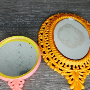 May include: Two hand mirrors with ornate frames. The larger mirror has a yellow frame with a detailed design. The smaller mirror has a pink frame with a yellow and pink trim.