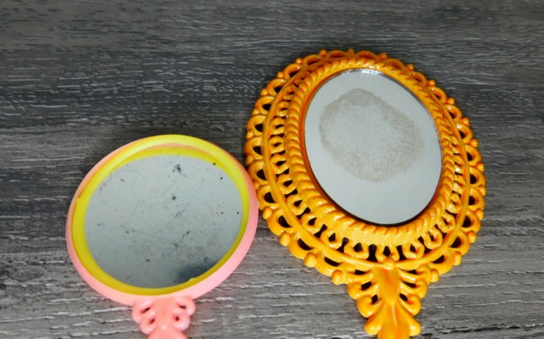 May include: Two hand mirrors with ornate frames. The larger mirror has a yellow frame with a detailed design. The smaller mirror has a pink frame with a yellow and pink trim.