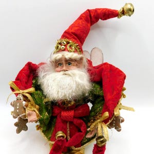 May include: A decorative Santa Claus figurine wearing a red hat with a gold bell at the tip, a green jacket with gold trim, and a red sash. The figure has a white beard, holds small gingerbread man ornaments, and features gold bells attached to the clothing.