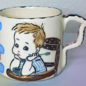 May include: Vintage ceramic child's mug with a whimsical illustration of a baby in a high chair. The mug features a white background with a black rim and handle, and a brown and gold base. The handle is decorated with blue and pink accents.