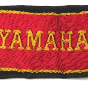 May include: A rectangular embroidered patch featuring the word YAMAHA in yellow thread on a red background. The patch is bordered by a thin yellow line and a thick black outer edge, creating a distinct contrast between the colors and the text.