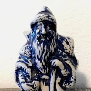 May include: A ceramic figurine of a wizard in a blue and white color scheme. The wizard is depicted with a long white beard, a pointed hat, and a staff. The figurine has a detailed, textured surface.