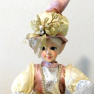 May include: A decorative elf figurine with a pink and gold outfit. The elf has a whimsical hat with a gold bell and a decorative hat with gold accents. The elf's sleeves are silver and the outfit has a sequined design.