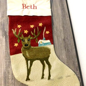 May include: A Christmas stocking featuring a cream-colored cuff with the name Beth embroidered in red. The main body is red with a textured green deer, yellow stars, and a small blue house with a white roof, all set against a light beige base.