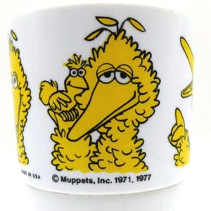 May include: White ceramic mug featuring Big Bird from Sesame Street. The mug has a yellow Big Bird graphic with a smaller bird. The mug also has the text "MADE IN USA" and "© Muppets, Inc. 1971, 1977".