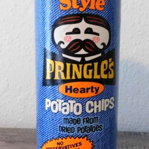 May include: A cylindrical can of Pringles potato chips. The can has a blue denim-like background with the words "Country Style" in orange. The Pringles logo features a cartoon character with a mustache. The can also states "Hearty Potato Chips" and "net wt. 4 1/2 oz."