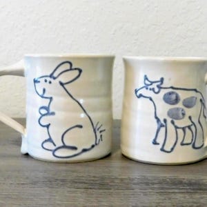 May include: Two ceramic mugs with cream-colored bodies and curved handles. One mug features a blue outline drawing of a rabbit, the other a cow. The mugs are on a wooden surface.
