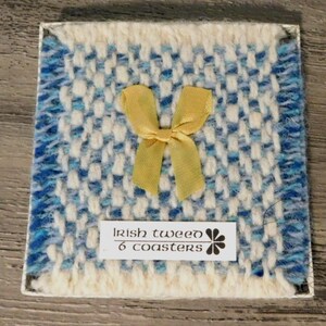May include: Square Irish tweed coaster with a woven blue and white pattern, a gold bow, and a label that reads "Irish tweed 6 coasters". The coaster has a white border.
