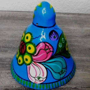 May include: A hand-painted ceramic bell with a vibrant floral design. The bell is primarily blue, with accents of pink, white, yellow, and green. The floral pattern is detailed and covers the bell's surface, creating a decorative piece.