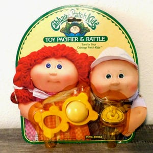 May include: A vintage Cabbage Patch Kids toy set featuring a girl and boy doll with pacifier and rattle. The dolls have rosy cheeks, blue eyes, and red and blonde hair. The packaging reads "TOY PACIFIER & RATTLE" and "Toys for Your Cabbage Patch Kids."