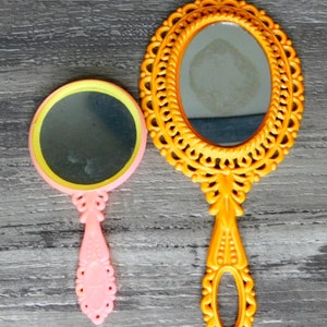 May include: Two hand mirrors, one with a yellow ornate frame and a clear glass mirror, and the other with a pink handle and a yellow and clear glass mirror.