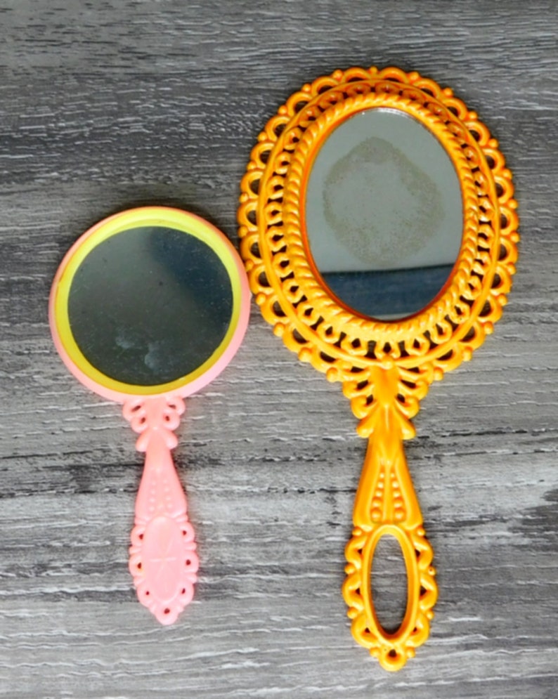 May include: Two hand mirrors, one with a yellow ornate frame and a clear glass mirror, and the other with a pink handle and a yellow and clear glass mirror.