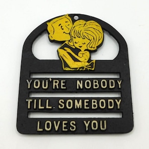 May include: Black plaque with yellow accents featuring a couple embracing. The text reads "YOU'RE NOBODY TILL SOMEBODY LOVES YOU". The plaque has a hole at the top for hanging.