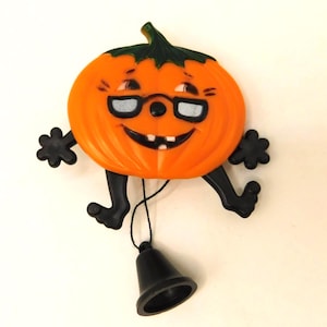 May include: A plastic orange pumpkin ornament with a green stem, black glasses, and a smiling face with two teeth. The figure has black arms and legs, and a black bell hangs from a thin black string attached to the bottom of the pumpkin body.