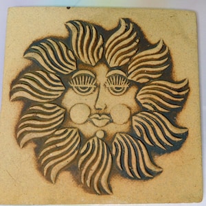 May include: A square, ceramic tile featuring a stylized sun face with a smiling expression. The sun has large, detailed eyes, rosy cheeks, and a full, smiling mouth. The sun's rays are depicted as wavy lines. The tile is tan with dark brown accents.