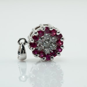 Può includere: Un ciondolo d'argento con un design rotondo che presenta un gruppo di piccoli diamanti bianchi circondati da un anello di rubini rossi.