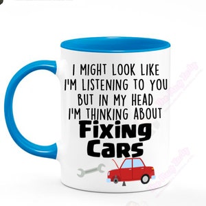 May include: White ceramic mug with a blue handle and interior. The mug features the text "I MIGHT LOOK LIKE I'M LISTENING TO YOU BUT IN MY HEAD I'M THINKING ABOUT Fixing Cars" with a red car and wrench graphic.