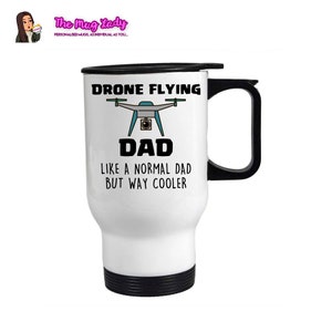Op de afbeelding: Witte reismok met een zwarte handgreep en deksel. De mok heeft de tekst "DRONE FLYING DAD LIKE A NORMAL DAD BUT WAY COOLER" met een drone-afbeelding. Het logo van The Mug Lady staat in de linkerbovenhoek.