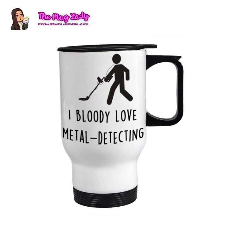 May include: White travel mug with a black lid and handle. The mug has a black silhouette of a person holding a metal detector with the text "I BLOODY LOVE METAL-DETECTING".