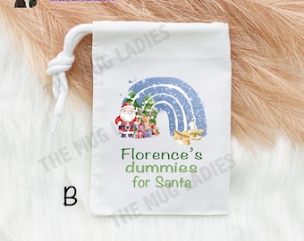 Op de afbeelding: Een witte trekzak met een blauw en wit regenboogontwerp. Onder de regenboog staan ​​de kerstman, een rendier en een slee. De tekst "Florence's dummies for Santa" staat onder de regenboog.