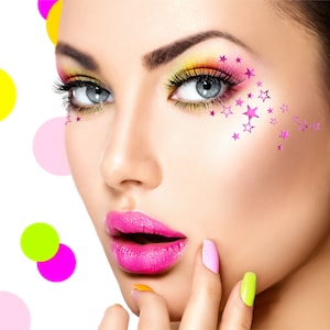 May include: A woman with pink eyeshadow and pink stars on her cheek. Her lips are painted a bright pink and she is wearing a light pink nail polish on one finger and a light green nail polish on another finger.