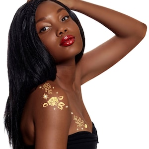 Może przedstawiać: Kobieta z długimi czarnymi warkoczami nosi czarną górę bez ramiączek i ma złote tymczasowe tatuaże róż i gwiazd na ramionach i klatce piersiowej.