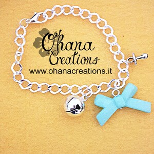 May include: A silver chain bracelet with a blue bow charm, a silver bell charm, and a silver pacifier charm. The bracelet has a clasp closure. The text "Ohana Creations" and "www.ohanacreations.it" is printed on a yellow background.