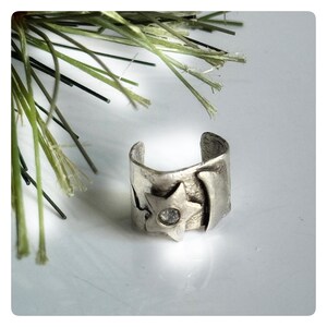 May include: A silver-toned cuff ring with a star and a clear stone. The ring has a textured surface and a cutout design. The ring is a unique piece of jewelry.