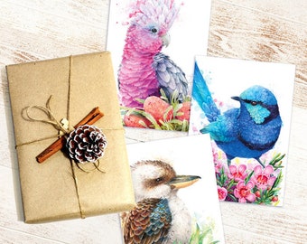 Può includere: Una collezione di biglietti di auguri ad acquerello con uccelli colorati, tra cui un cacatua rosa, uno scricciolo blu e un kookaburra. È presente anche un regalo avvolto con un bastoncino di cannella e una pigna, insieme a decorazioni natalizie.