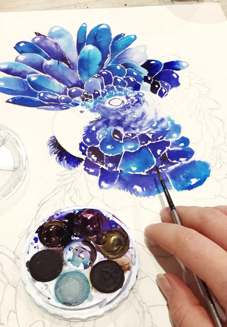 Puede incluir: Un primer plano de una pintura de acuarela de un p&aacute;jaro azul con una cresta azul y morada. El artista est&aacute; usando un pincel peque&ntilde;o para agregar detalles a la pintura. Una paleta de acuarela con varios tonos de azul, morado y negro es visible en primer plano.