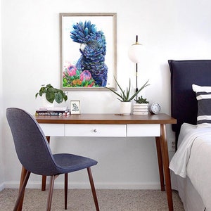 Puede incluir: Una pintura de acuarela de un cacat&uacute;a negro con una cresta azul y morada, rodeado de flores de protea rosas. La pintura est&aacute; enmarcada y colgada en una pared blanca sobre un escritorio con una silla.