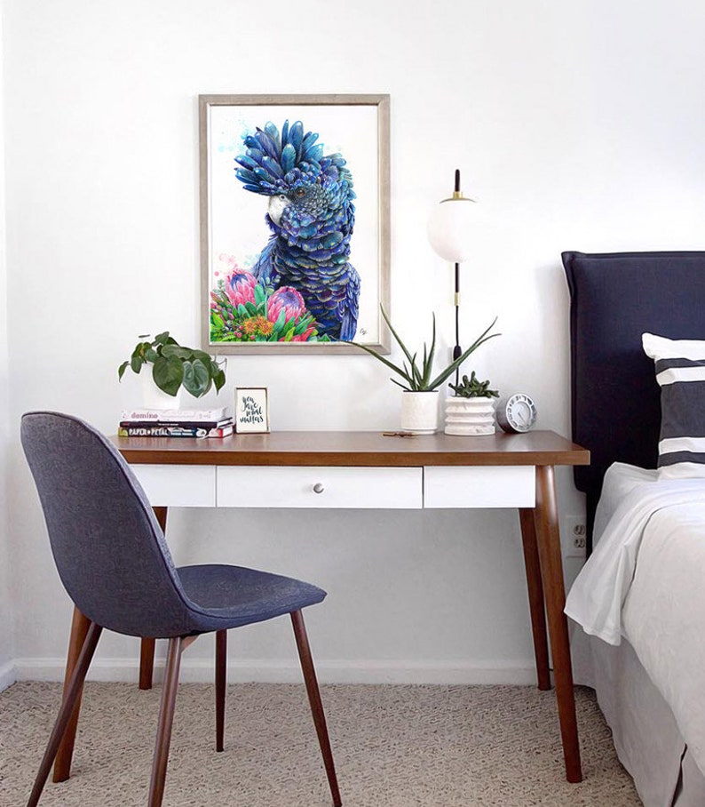 Puede incluir: Una pintura de acuarela de un cacat&uacute;a negro con una cresta azul y morada, rodeado de flores de protea rosas. La pintura est&aacute; enmarcada y colgada en una pared blanca sobre un escritorio con una silla.