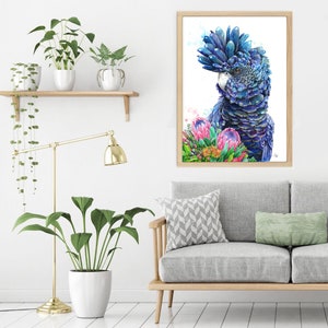 Puede incluir: Una pintura de acuarela de un cacat&uacute;a azul y negro con una flor de protea rosa y verde. La pintura est&aacute; enmarcada y colgada en una pared blanca en un ambiente de sala de estar.