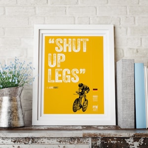 以下が含まれることがあります： 黄色の背景に白い文字で「SHUT UP LEGS」と書かれた白い額縁のポスター。ポスターには、白黒のサイクリストの画像と、白い文字で「JENS VOIGT」と「PTW ART」と書かれている。