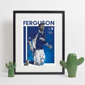 May include: Framed print featuring a soccer player in a blue and white uniform, kneeling on a blue background. The print includes the name "FERGUSON" and the number "9". The artwork is displayed with two decorative cactus figurines.