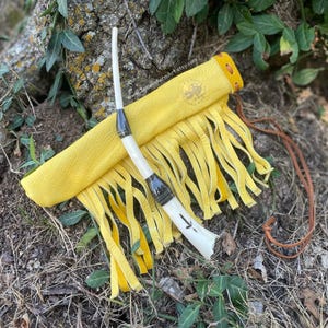 May include: A handcrafted, yellow leather sheath with fringe, holding a bone-handled tool with black accents. The tool and sheath rest on a bed of earth and foliage. A brown leather cord is also present.