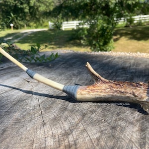 May include: A handcrafted antler pipe with a light-colored stem and a brown, textured antler bowl. The stem is wrapped with blue thread. The pipe rests on a weathered wooden surface.