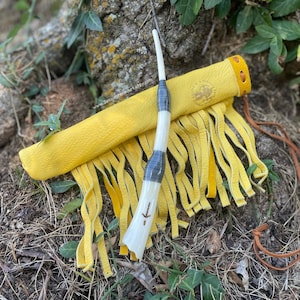 May include: A handcrafted, yellow leather quiver with fringe, a white and gray wand, and a brown leather strap. The quiver has a sun-like emblem. The wand has a carved arrow design. The items are set on a bed of dry grass and leaves.
