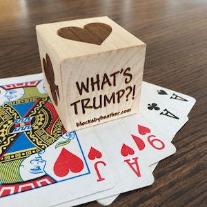 May include: A wooden block with a heart cutout and the text "What's Trump?!" The block is sitting on top of a deck of playing cards, including the Ace of Spades and the Ace of Hearts.
