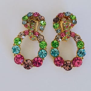 May include: A pair of clip-on earrings with a gold-tone metal setting. Each earring features a circular design adorned with colorful, round rhinestones in shades of pink, green, blue, and brown. The earrings are set against a white background.