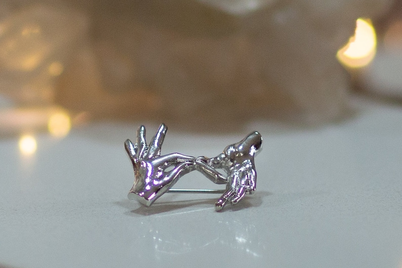 May include: Silver brooch depicting two hands clasped together, representing love and unity.