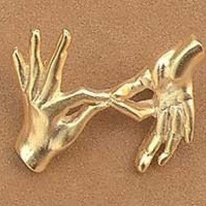 May include: A gold-colored metal brooch depicting two hands clasped together. The brooch has a polished finish and is likely made of brass or a similar alloy. The hands are detailed with visible fingers and a realistic form.
