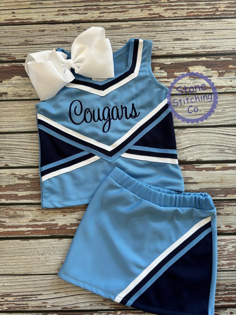 May include: A light blue cheerleading uniform set with a matching skirt and a large white bow. The top features the word "Cougars" in black script. The uniform has navy blue and white accents.