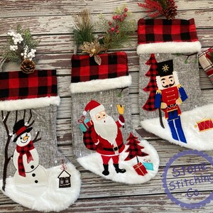 May include: Three grey Christmas stockings with white fur trim and red and black plaid cuffs. The stockings are decorated with embroidered images of a snowman, Santa Claus, and a nutcracker.