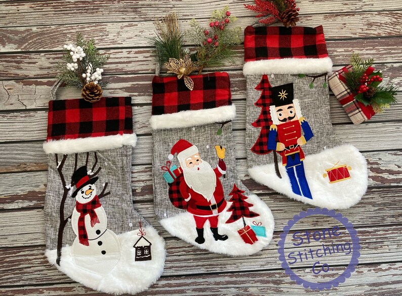 May include: Three grey Christmas stockings with white fur trim and red and black plaid cuffs. The stockings are decorated with embroidered images of a snowman, Santa Claus, and a nutcracker.