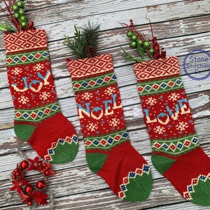 May include: Three red knitted Christmas stockings with green, blue, and white patterns. The stockings have the words "JOY", "NOEL", and "LOVE" embroidered on them.