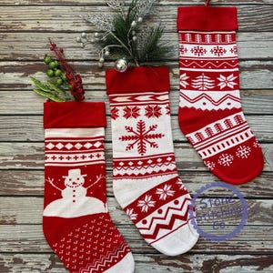 May include: Three red and white Christmas stockings with different patterns. The stocking on the left has a snowman design, the middle stocking has a snowflake design, and the stocking on the right has a geometric pattern. The stockings are hanging on a wooden background.