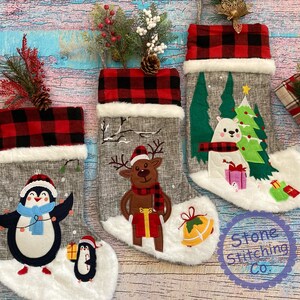 May include: Three grey and white Christmas stockings with red and black checked cuffs. The stockings feature embroidered designs of a penguin, a reindeer, and a polar bear. The stockings are decorated with faux fur and are ready to be filled with gifts.
