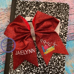 May include: Red glitter hair bow with a white rhinestone centre. The bow has the name "JAELYNN" embroidered on one side and "PRE-K" with a red apple and books on the other side.