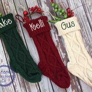 May include: Three knitted Christmas stockings with personalized names: Jake, Koelli, and Gus. The stockings are green, red, and white.